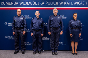 Grafika przedstawia trzech policjantów i policjantkę stojących na tle granatowego baneru z białym napisem komenda Wojewódzka Policji w Katowicach.