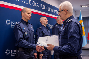 Grafika przedstawia Komendanta Wojewódzkiego Policji w Katowicach gratulującego awansu policjantowi stojącemu na przeciw niego.