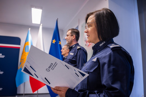 Na grafice na pierwszym planie widać policjantkę stojącą bokiem trzymającą w rękach r biała teczkę z napisem "Policja Komenda Wojewódzka Policji w Katowicach". na drugim planie widoczni są stojący za nią dwaj policjanci.