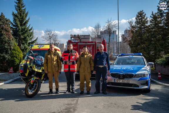 Na zdjęciu Zastępca Naczelnika Wydziału Ruchu Drogowego Komendy Wojewódzkiej Policji w Katowicach, Dyrektor Wojewódzkiego Pogotowia Ratunkowego, Rzecznik Prasowy Śląskiego Komendanta Wojewódzkiego Państwowej Straży Pożarnej oraz Naczelnik Wydziału Operacyjno-Szkoleniowego Komendy Miejskiej Straży Pożarnej w Katowicach. Za nimi radiowóz, wóz strażacki, karetka pogotowia i motor