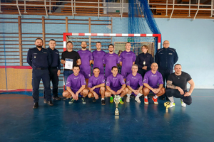 Zdjęcie zwycięskiej drużyny wra zz pucharem i dyplomem na tle bramki wraz z naczelnikami z Komendy Wojewódzkiej Policji w Katowicach.