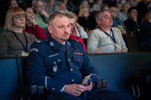 Zdjęcie przedstawia zbliżenie na policjanta siedzącego na widowni