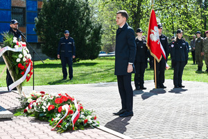 Mężczyzna w stroju cywilnym składa kwiaty pod pomnikiem, w tle sztandar klasy mundurowej