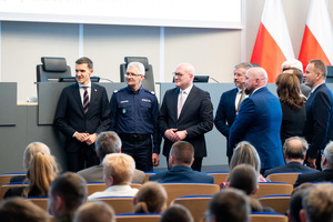 Generał Policji stoi w auli z mężczyznami w garniturach, inne osoby siedzą na krzesłach