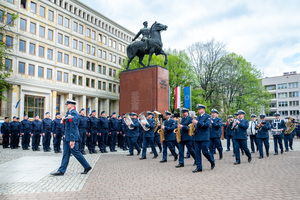 Umundurowani policjanci stoją przy pomniku na placu, przed nimi przechodzi policyjna orkiestra