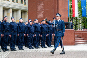 Umundurowani policjanci stoją przy pomniku na placu, przed nimi przechodzi dowódca uroczystości, trzyma w ręku szablę
