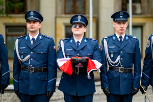 Trzech policjantów w mundurach galowych stoi w szeregu, policjant stojący w środku w rękach trzyma flagę Polski