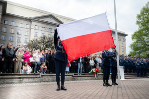 Trzech policjantów w mundurach galowych wciąga na maszt flagę Polski, w tle grupa osób w cywilnych ubraniach, obok nich policjanci w mundurach galowych