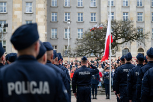 Flaga Polski jest wciągana na maszt przez policjantów, maszt stoi na placu, na którym policjanci w mundurach galowych stoją w szeregach, w tle grupa osób w ubraniach cywilnych