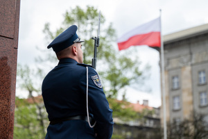Policjant w mundurze galowym pełni wartę przy pomniku, w rękach trzyma karabin z bagnetem, w tle biało czerwona flaga na maszcie