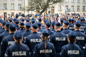 Policjanci w mundurach ćwiczebnych stoją w kilku szeregach w tle widać grupę osób w ubraniach cywilnych