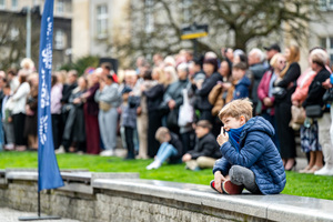 Kilkuletni chłopiec siedzi na murku, w tle grupa osób w ubraniach cywilnych