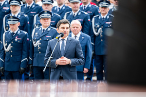Przy mikrofonie stoi mężczyzna w garniturze, w tle policjanci w mundurach galowych