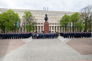 Przed pomnikiem policjanci i policjantki w mundurach ćwiczebnych, policjanci w mundurach galowych oraz osoby w ubraniach cywilnych stoją w kilku szeregach