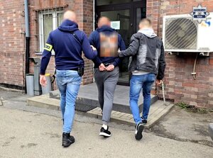 nieumundurowani policjanci prowadzą osobę, znajduję się przed budynkiem komisariatu Policji