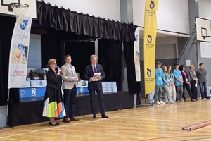 Zdjęcie przedstawia dwóch mężczyzn oraz kobietę która w ręce trzyma mikrofon w tle po prawej stronie stoją uczestnicy konkursu