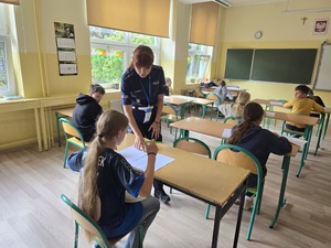 Zdjęcie przedstawia uczniów rozwiązujących test wiedzy oraz policjanta tłumaczącego polecenia
