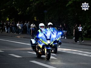 Zdjęcie przedstawia dwóch policjantów jadących na motocyklach. W ich tle znajdują się ludzie idący po chodniku.
