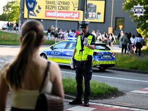 Na zdjęciu widoczny jest policjant kierujący ruchem. W jego tle znajduje się radiowóz oraz idący ludzie.
