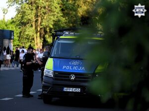 Na zdjęciu widocznych jest dwóch policjantów przed radiowozem. W ich tle widać ludzi.