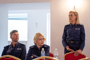 Na zdjęciu widocznych jest trzech policjantów. Jedna z nich stoi i przemawia.