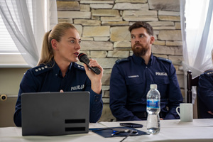Na zdjęciu widocznych jest dwóch policjantów, którzy zasiadają przy stole. Jeden z nich trzyma w ręku mikrofon, do którego mówi.