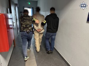 Na zdjęciu znajduje się dwóch nieumundurowanych policjantów, którzy prowadzą zatrzymanego mężczyznę.