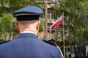 Na pierwszym planie zdjęcia widoczny jest odwrócony plecami policjant, a w jego tle widoczna jest flaga Polski i krzyż
