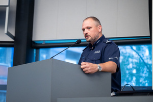 Na zdjęciu widoczny jest policjant stojący za mównicą.