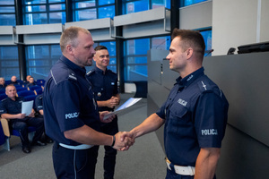Zdjęcie przedstawia policjantów podających sobie dłoń, a w ich tle widoczna jest grupa policjantów, która siedzi.