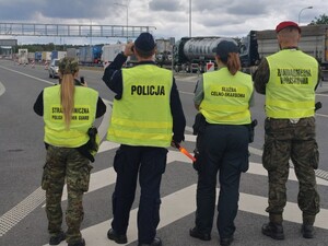 na zdjęciu widoczni stojący na drodze tyłem funkcjonariusze w zielonych kamizelkach żandarmerii wojskowej, służby celno skarbowej , straży granicznej i policjant  w oddali widoczne pojazdy ciężarowe stojące w kierunku granicy