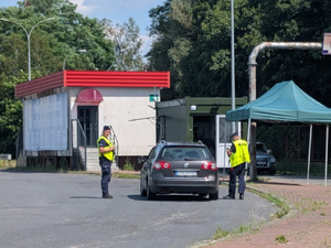 na zdjęciu widoczni stojący na drodze w zielonych kamizelkach policjanci, dokonujący kontroli samochodu osobowego w oddali zabudowania