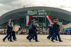 zdjęcie przedstawia maszerujących policjantów orkiestry na tle areny pre zero