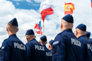 zdjęcie przedstawia policjantów stojących w kolumnie a pomiędzy nimi widok na powiewające flagi polski, hiszpanii austrii i czech
