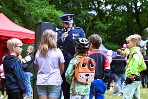 zdjęcie przedstawia policjanta mówiącego przez mikrofon a obok grupa dzieci