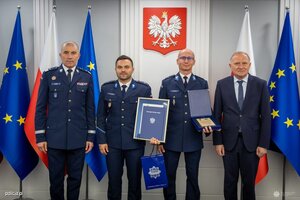 na zdjęciu widoczni stojący w auli trzej policjanci i mężczyzna w ciemnym garniturze za osobami flagi polski i unii europejskiej a w oddali na ścianie zawieszone godło polski