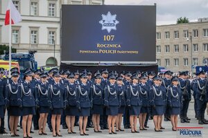 na zdjęciu widoczni stojący w szeregu umundurowani policjanci w tle zabudowania oraz ekran z napisem sto siódma rocznica powstania policji państwowej nad nim gwiazda policyjna z napisem policja