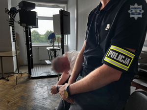 na zdjęciu widoczny na pierwszym planie  w pokoju policjant w granatowej koszulce  z opaską z napisem policja założoną na ramieniu, za nim siedzi mężczyzna z założonymi na ręce kajdankami, w oddali w pokoju widoczne stanowisko do wykonywania zdjęć sygnalitycznych