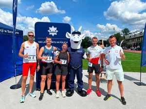 na zdjęciu widoczni stojący  na boisku  czterej mężczyźni i jedna kobieta w strojach sportowych, pomiędzy nimi stoi maskotka policyjna, biały koziołek w niebieskim mundurze z napisem policja, za osobami niebieski baner z logo i gwiazdą Policja oraz napisem 100 lat sportu w policji, w oddali  bieżnia sportowa i zabudowania.
