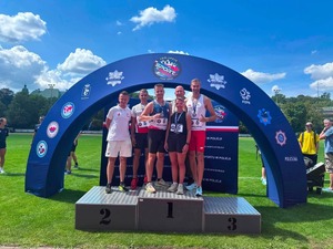 na zdjęciu widoczni stojący na podeście usytuowanym na trawiastym boisku 5 mężczyzn i 1 kobieta w strojach sportowych, osoby te mają zawieszone okolicznościowe medale, za osobami widoczny baner koloru niebieskiego z napisem 100-lecie Sport w Policji, na dalszym planie widoczne drzewa i dachy zabudowań