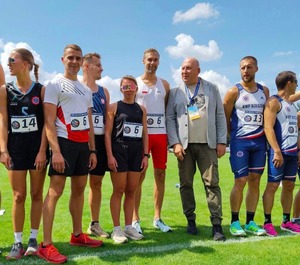 na zdjęciu widoczni stojący na trawiastym boisku 5 mężczyzn i 2 kobiety  w strojach sportowych, pomiędzy nimi mężczyzna w brązowych spodniach i jasnej marynarce.