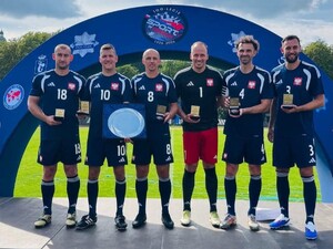 na zdjęciu widoczni stojący na podeście usytuowanym na trawiastym boisku 6 mężczyzn w strojach sportowych koloru niebieskiego , którzy trzymają w rękach okolicznościowe medale, a jeden z nich również srebrną tacę , za mężczyznami widoczny baner koloru niebieskiego z napisem 100-lecie Sport w Policji
