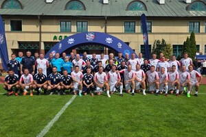 na zdjęciu widoczni stojący na murawie boiska mężczyźni z dwóch drużyn  w niebieskich i biało czerwonych strojach sportowych, niebieski baner za stojącymi osobami posiada w górnej części napis 100- lecie Sport w Policji