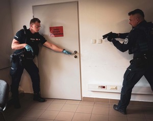 na zdjęciu widoczny pokój oraz zamknięte drzwi z napisem zakaz wstępu serwerownia, po lewej stronie drzwi widoczny umundurowany policjant trzymający w prawej ręce pistolet, a lewą ręką trzyma klamkę , po prawej stronie drzwi widoczny drugi umundurowany policjant trzymający w rękach pistolet skierowany w stronę drzwi