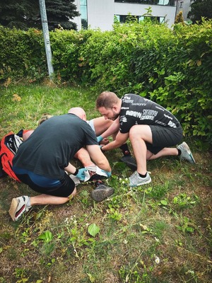 na zdjęciu widoczne ujęcie na trawnik ogrodzony żywopłotem, pośrodku trawnika widoczni dwaj mężczyźni w strojach sportowych udzielający pomocy medycznej leżącej osobie, obok nich czerwona torba medyczna, w oddali widoczne zabudowania.