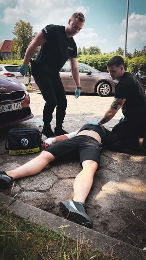 na zdjęciu widoczne ujęcie na drogę na której przed zaparkowanym pojazdem na ziemi leży na plecach osoba w krótkich czarnych spodenkach i czarnej koszulce z zabandażowaną prawą dłonią i prawym łokciem, obok leżącej osoby na ziemi widoczna torba medyczna z napisem Policja, przy mężczyźnie widoczni dwaj  policjanci udzielający leżącemu pomocy medycznej