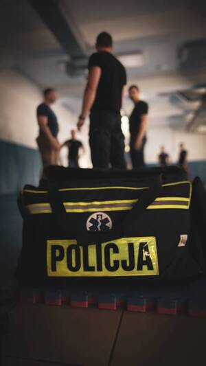 na zdjęciu widoczna na pierwszym planie torba medyczna z napisem Policja leżąca na macie sportowej w salce sportowej, na drugim planie 6 osób ćwiczących na macie