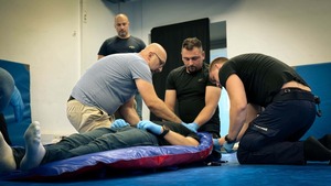 na zdjęciu widoczne ujęcie na salę sportową w centralnym kadrze widoczna osoba leżąca na ułożonych na podłodze noszach, leżącej osobie trzej mężczyźni udzielają pomocy medycznej, na drugim planie widoczny stojący mężczyzna  który przygląda się wykonywanym czynnościom