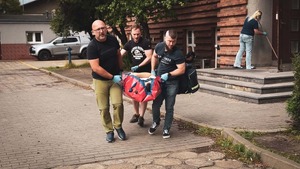 na zdjęciu widoczna część ulicy i chodnik przed wejściem do budynku , w centralnej części zdjęcia widoczni trzej mężczyźni niosący  leżącą na czerwonych noszach osobę , w oddali zaparkowany pojazd koloru szarego