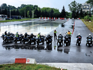 na zdjęciu widoczni usytuowani na placu manewrowym, w szeregu policjanci w czarnych kombinezonach motocyklowych i białych kaskach, siedzący na oznakowanych motocyklach służbowych z napisem policja obok nich instruktorzy policyjni